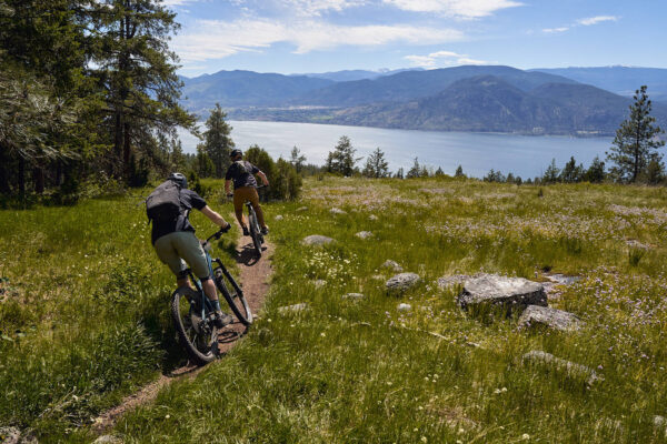 2 mountain bikers on singletrack with a beautiful lake and mountain scene in the background