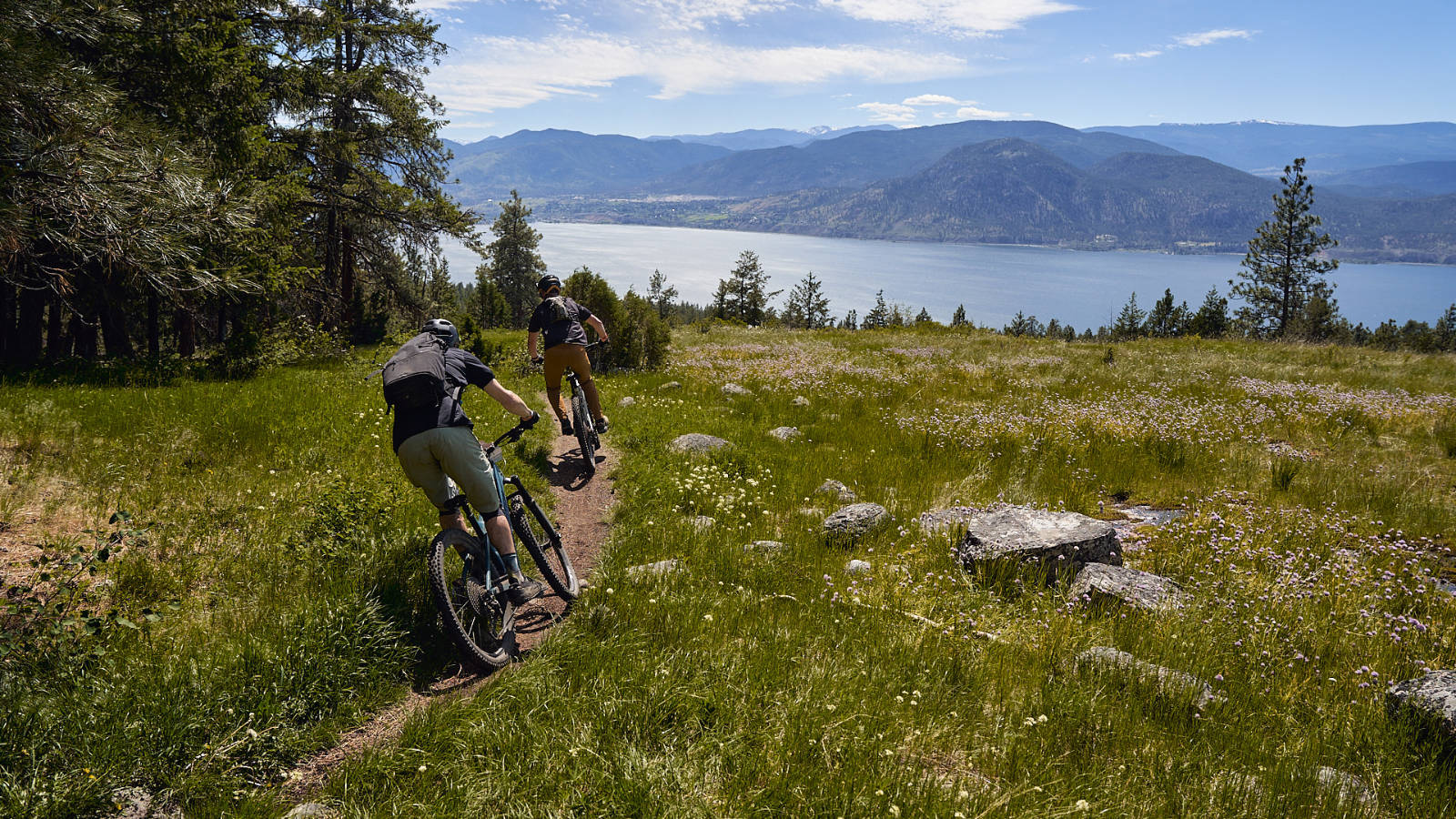 2 mountain bikers on singletrack with a beautiful lake and mountain scene in the background